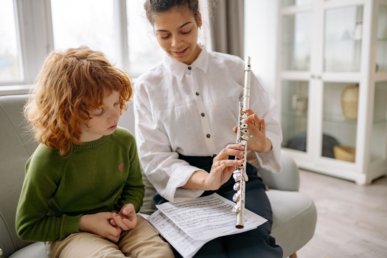 A young boy learns to play the flute with a teacher at home, surrounded by music sheets.